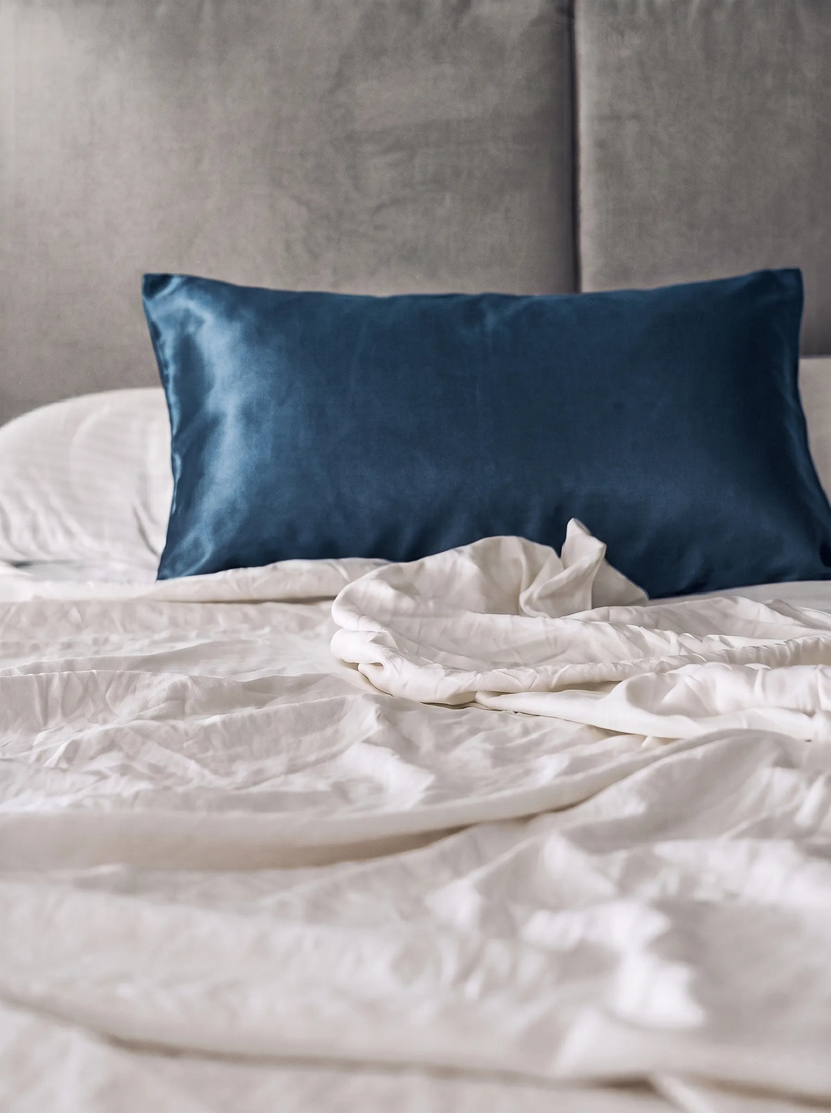 Taie d'oreiller en soie 19 mommes bleu paon sur lit blanc avec fond gris.