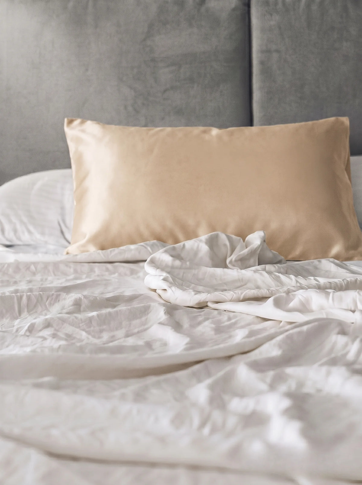 Taie d'oreiller en soie 19 mommes champagne sur lit blanc avec fond gris.