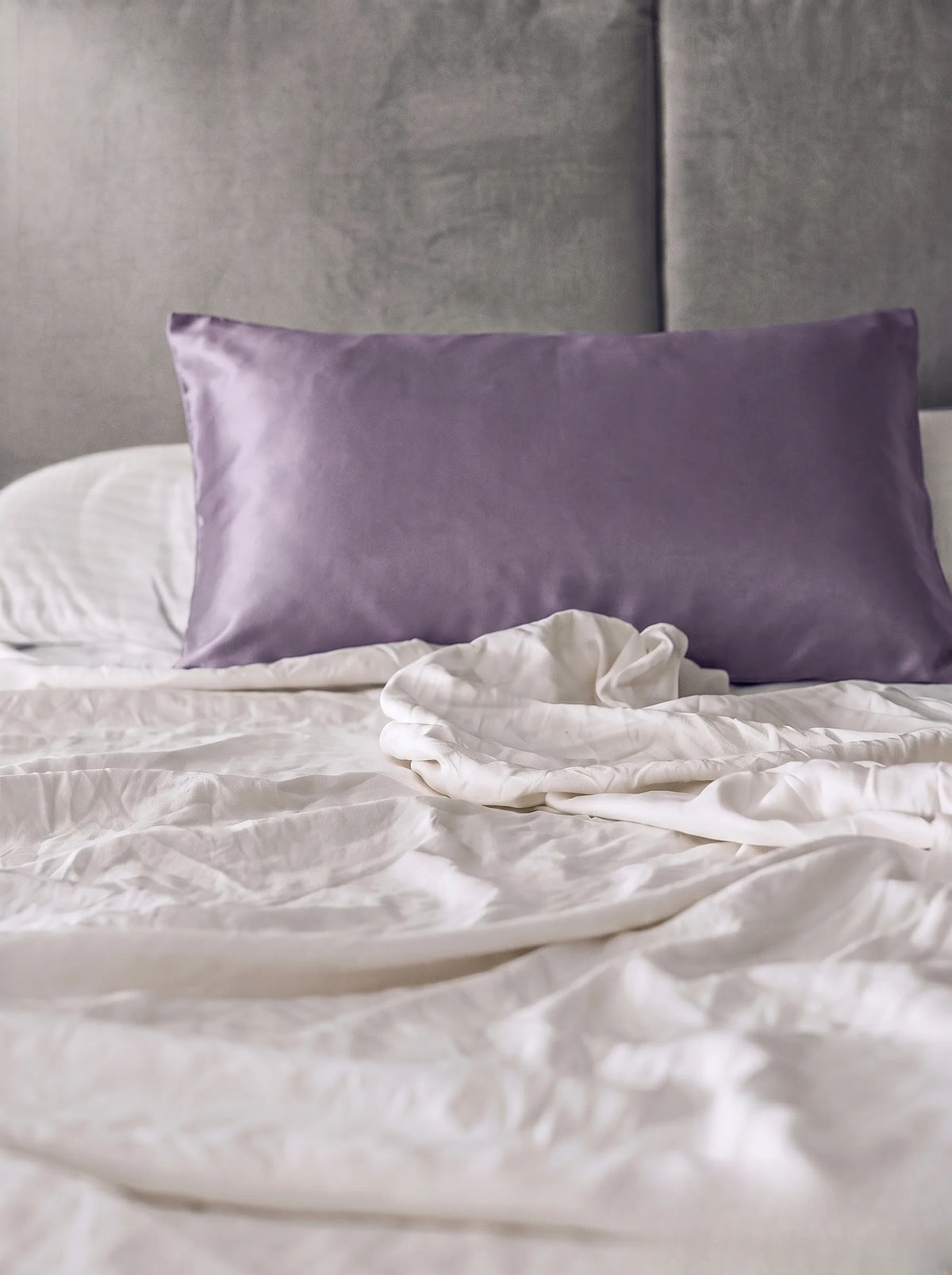 Taie d'oreiller en soie 19 mommes lavande sur lit blanc avec fond gris.